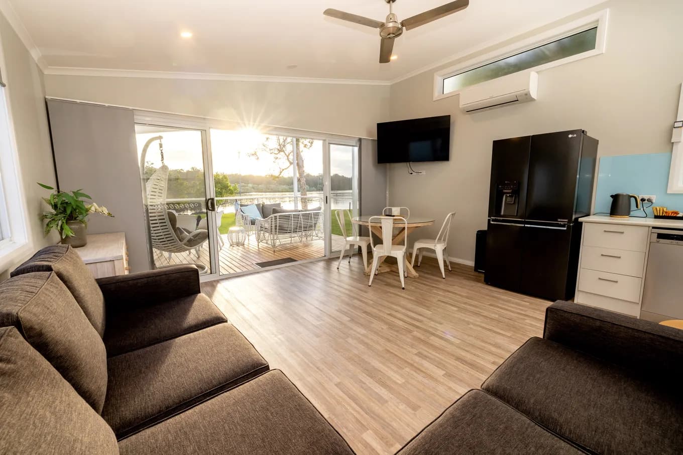 Open-plan living area in the Luxury Riverview Villa Two Bedroom: two brown fabric lounges on timber-look floors, a ceiling fan and split system overhead, a wall-mounted TV, and sliding doors opening to the riverfront deck.