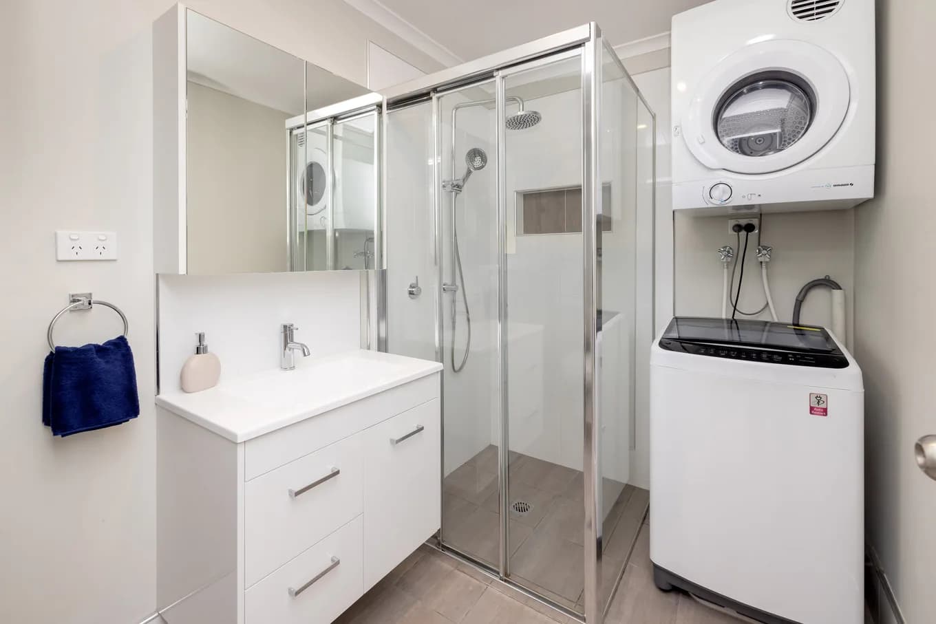 Bathroom in the two bedroom villa, with a glass-screened shower, a white vanity with a mirror, and a stacked washing machine and dryer beside it.