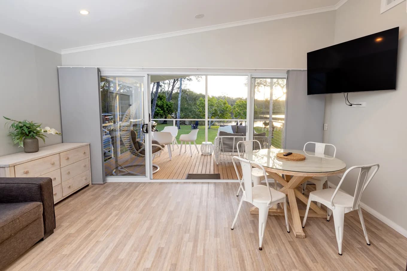 Living and dining space in the two bedroom villa: a round timber table with white metal chairs, a wall-mounted TV, a timber sideboard, and sliding doors onto the deck.