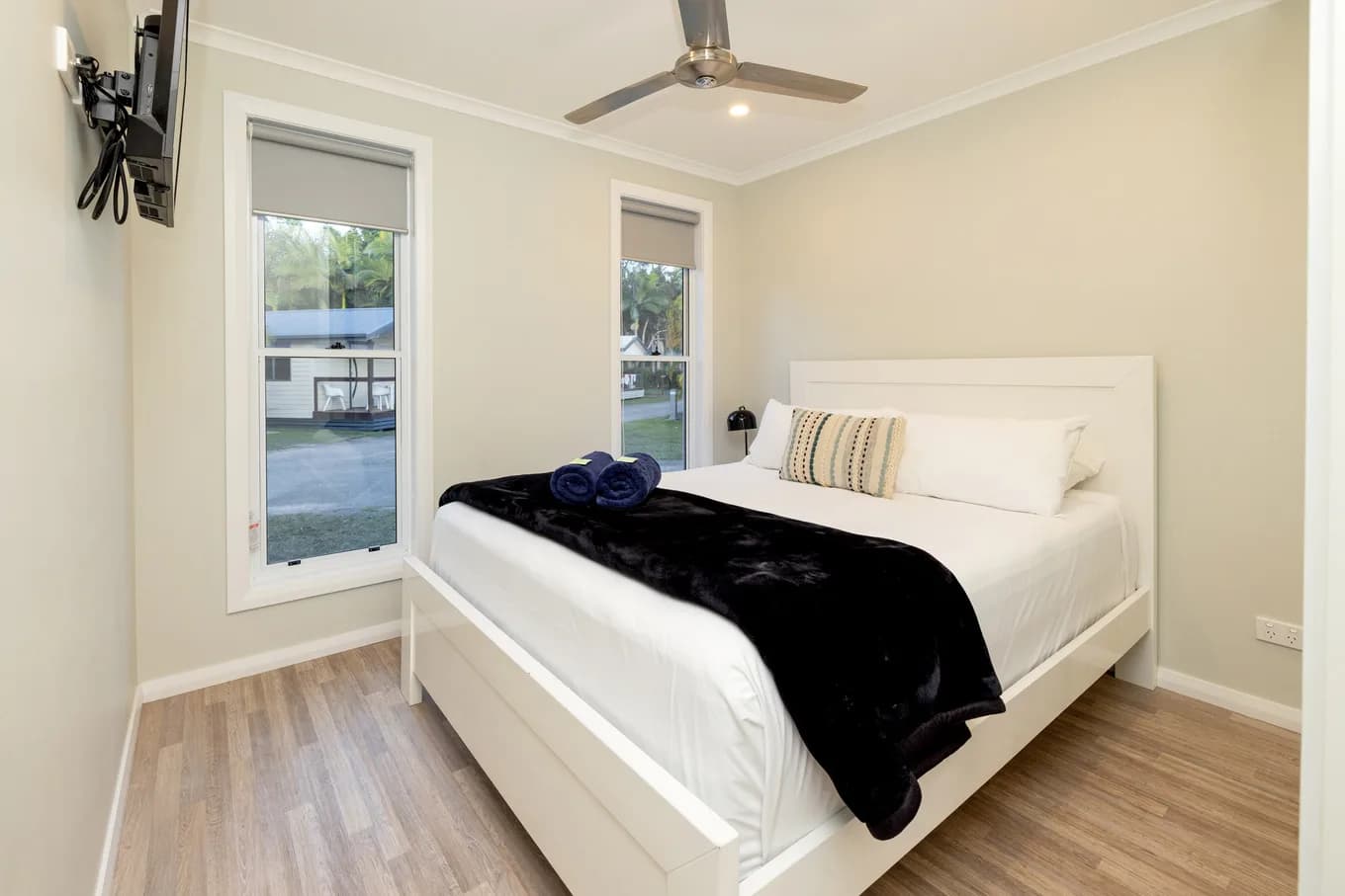 Queen bedroom in the two bedroom villa, made up in white linen with a black throw and rolled towels, a wall-mounted fan and windows on two sides.