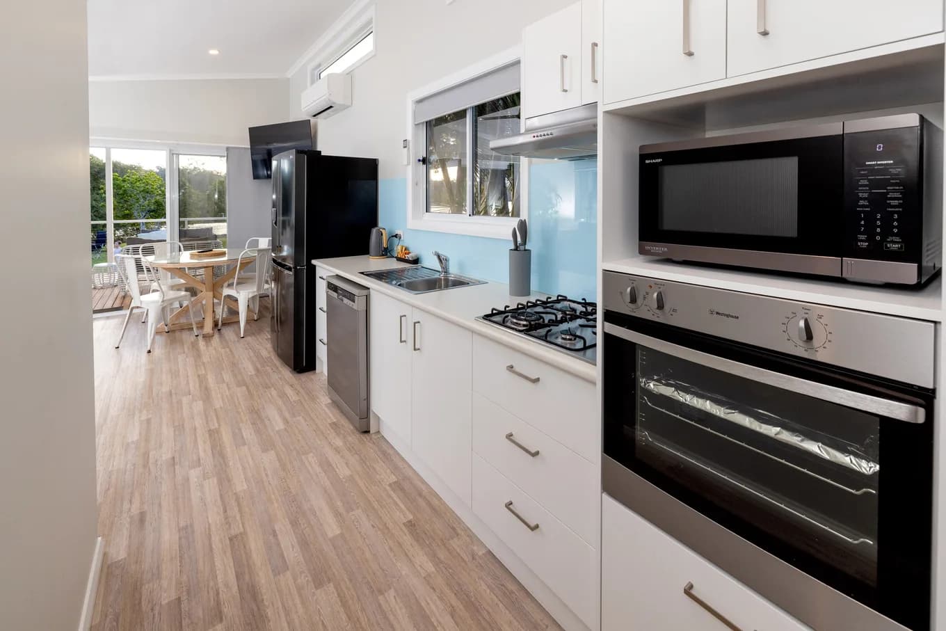 Galley kitchen in the two bedroom villa: a wall oven and microwave, gas cooktop, dishwasher and a black fridge along white cabinetry, with the dining area beyond.