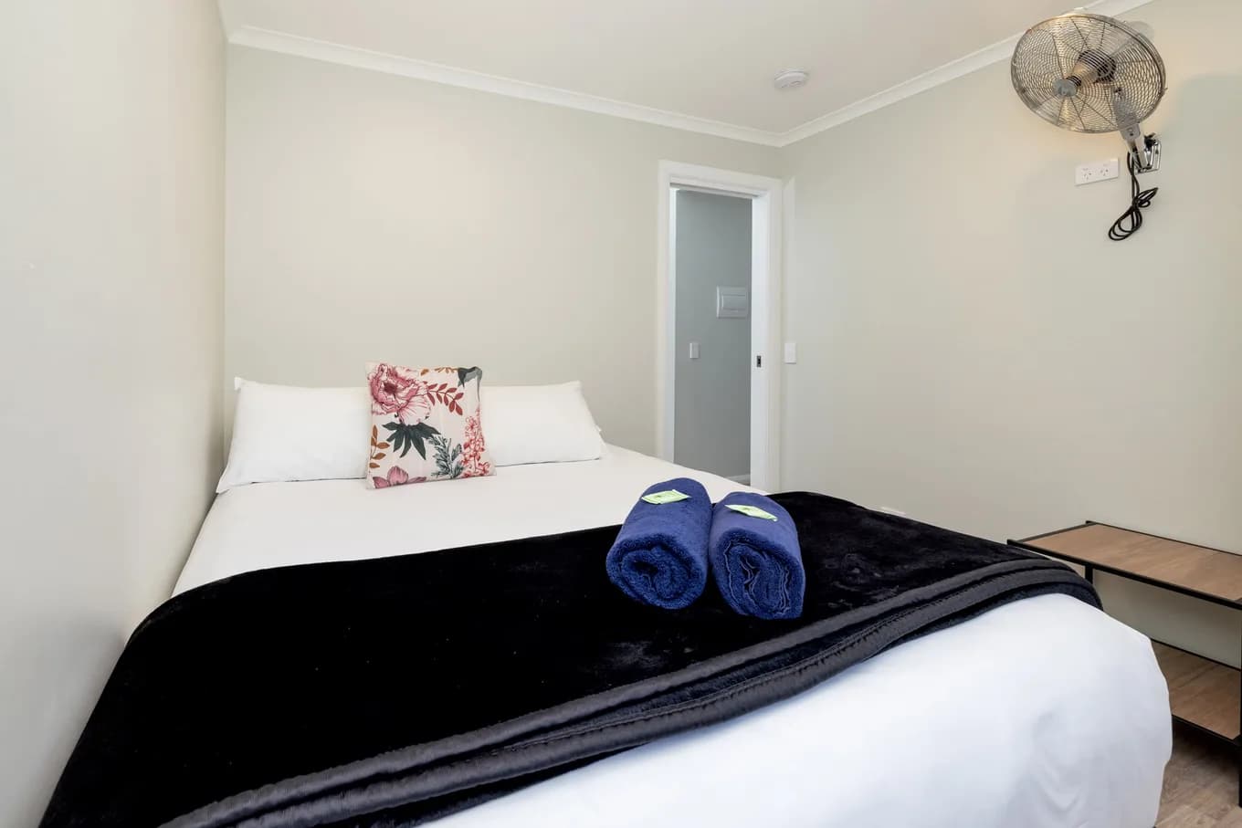Second bedroom in the two bedroom villa, with a bed made up in white under a black throw, a floral cushion, a wall-mounted fan and a door through to the hallway.