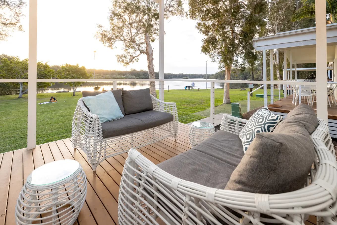 The villa covered deck at golden hour, with woven outdoor lounges and cushions looking across the lawn to the Wooli Wooli River.